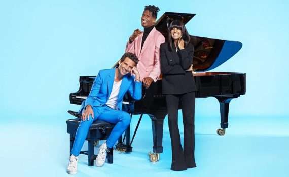 The three presenters sitting and standing around a grand piano.